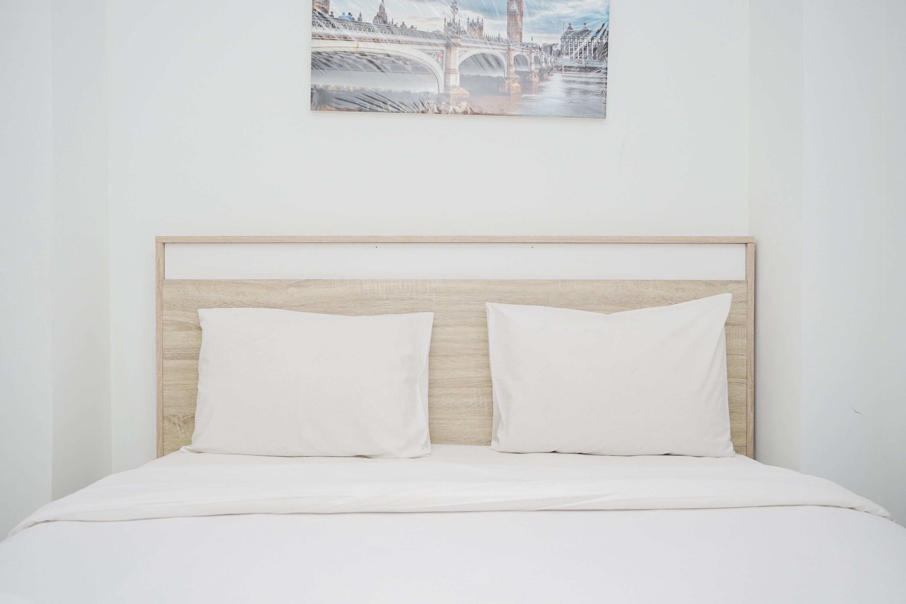 A bed with a wooden headboard, white bedding, and a cityscape painting above.