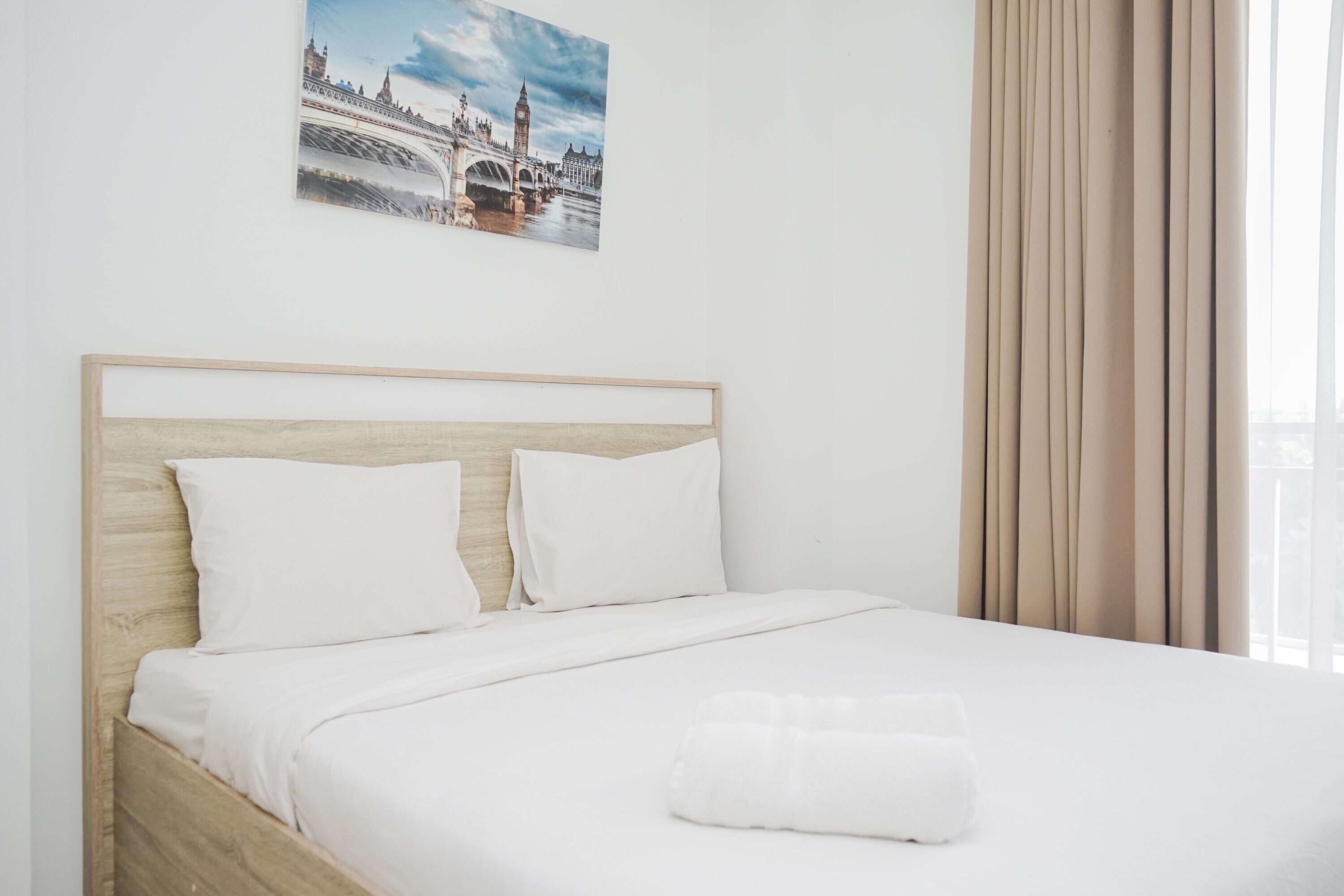 A neatly made bed with white linens and a rolled towel, a wooden headboard, and a framed picture of a bridge on the wall.