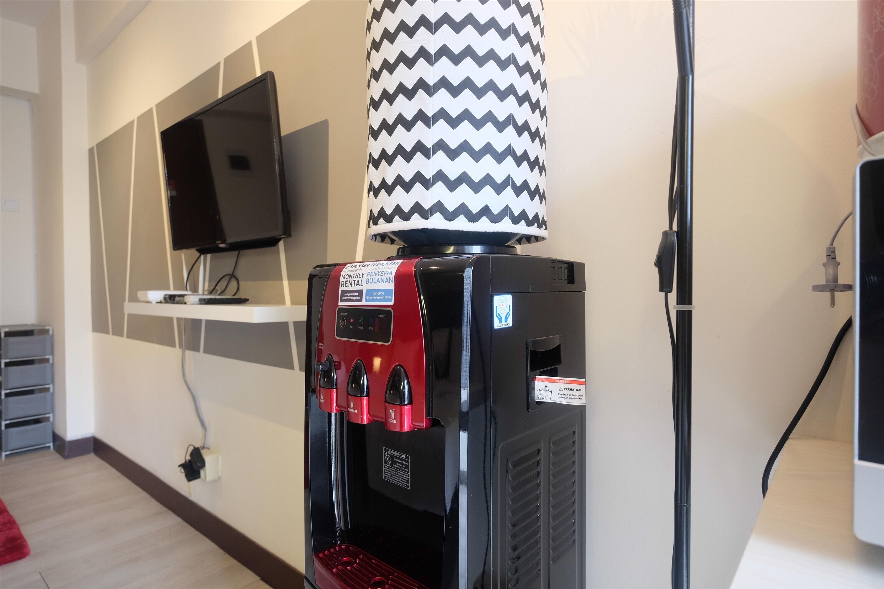A water dispenser with a red control panel, a television mounted on the wall, and a patterned lamp.