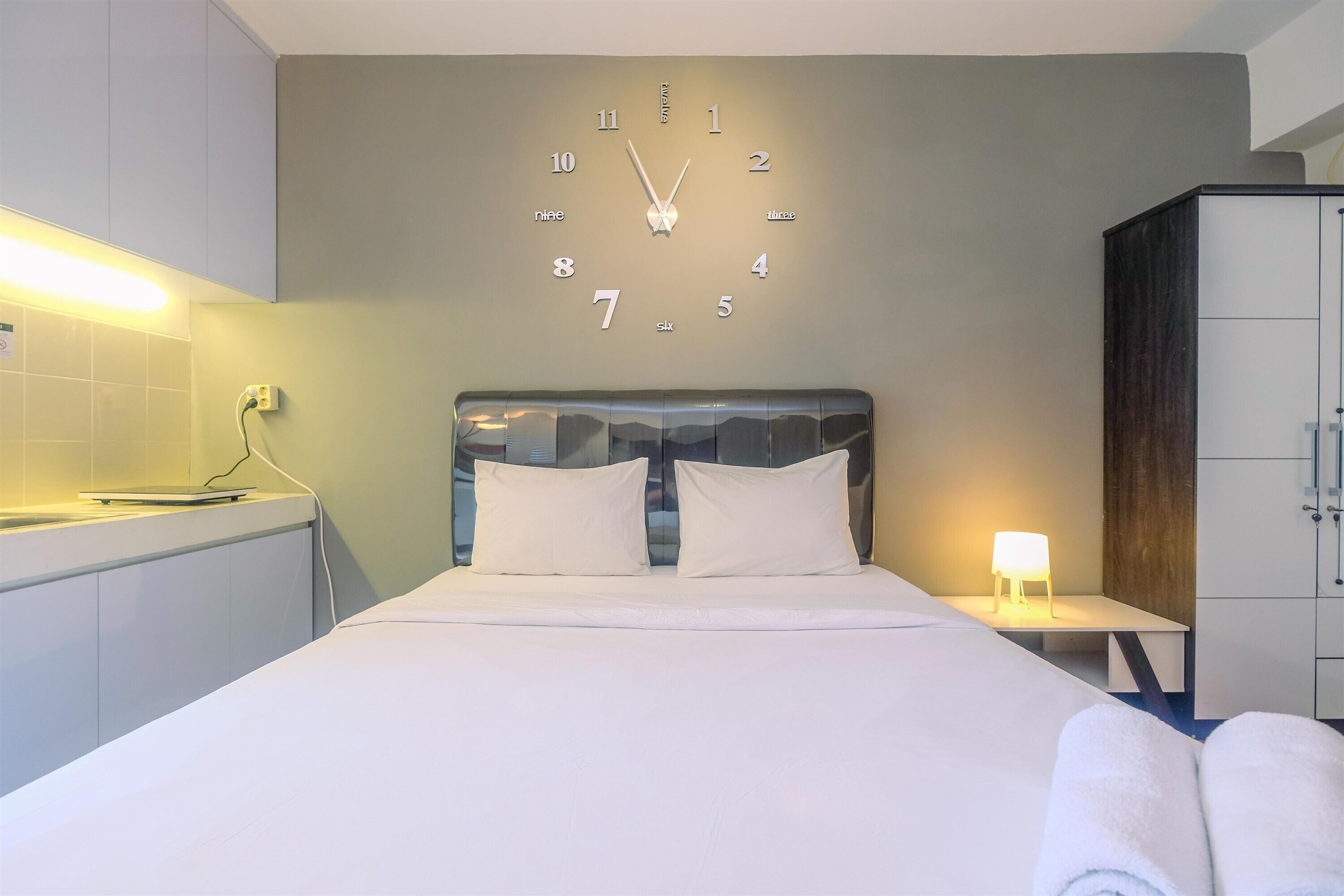 A hotel room with a bed, a wall clock, a bedside table with a lamp, and a wardrobe.