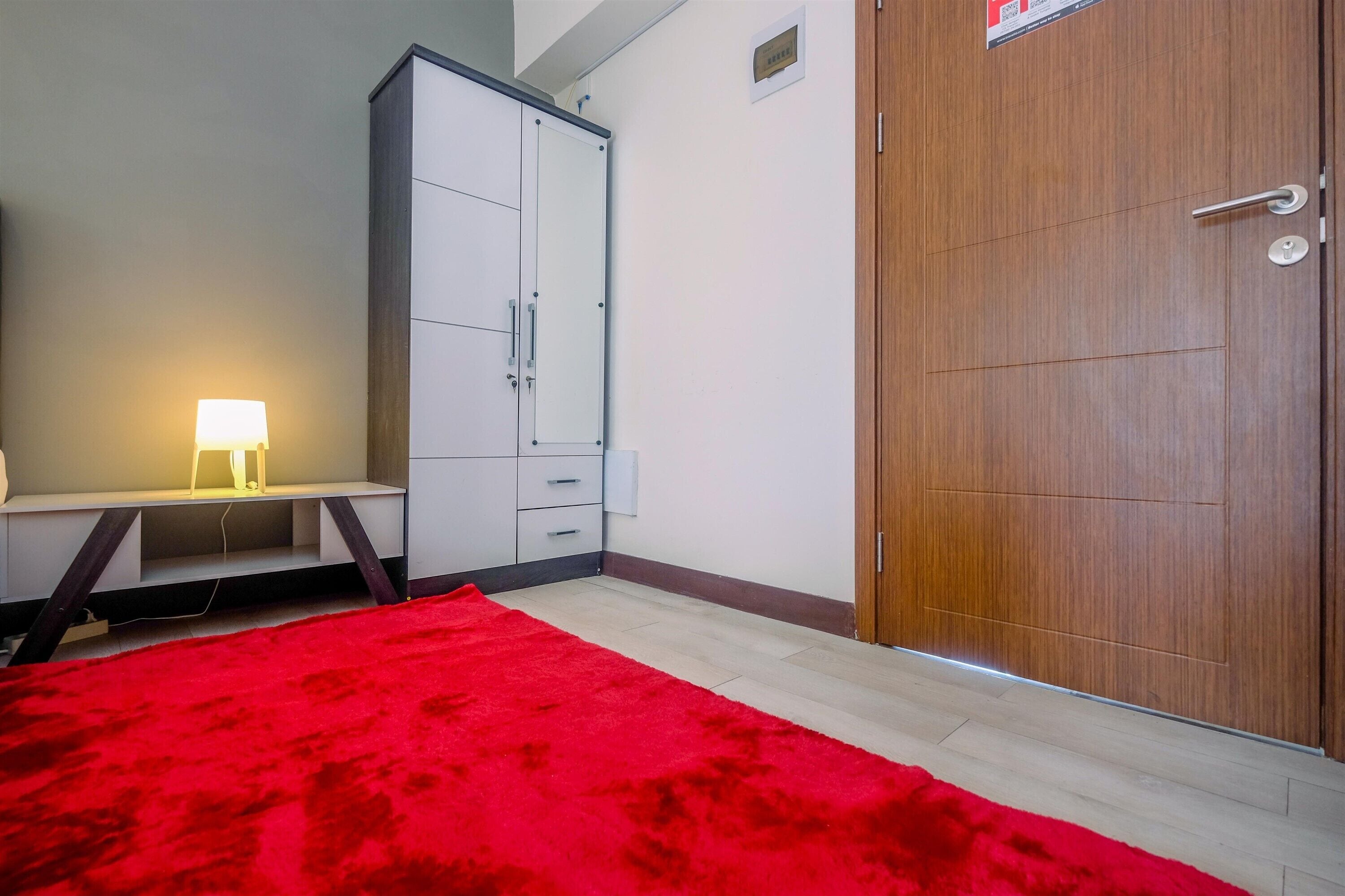 Quarto com carpete vermelho, escrivaninha com luminária, guarda-roupa e porta de madeira.