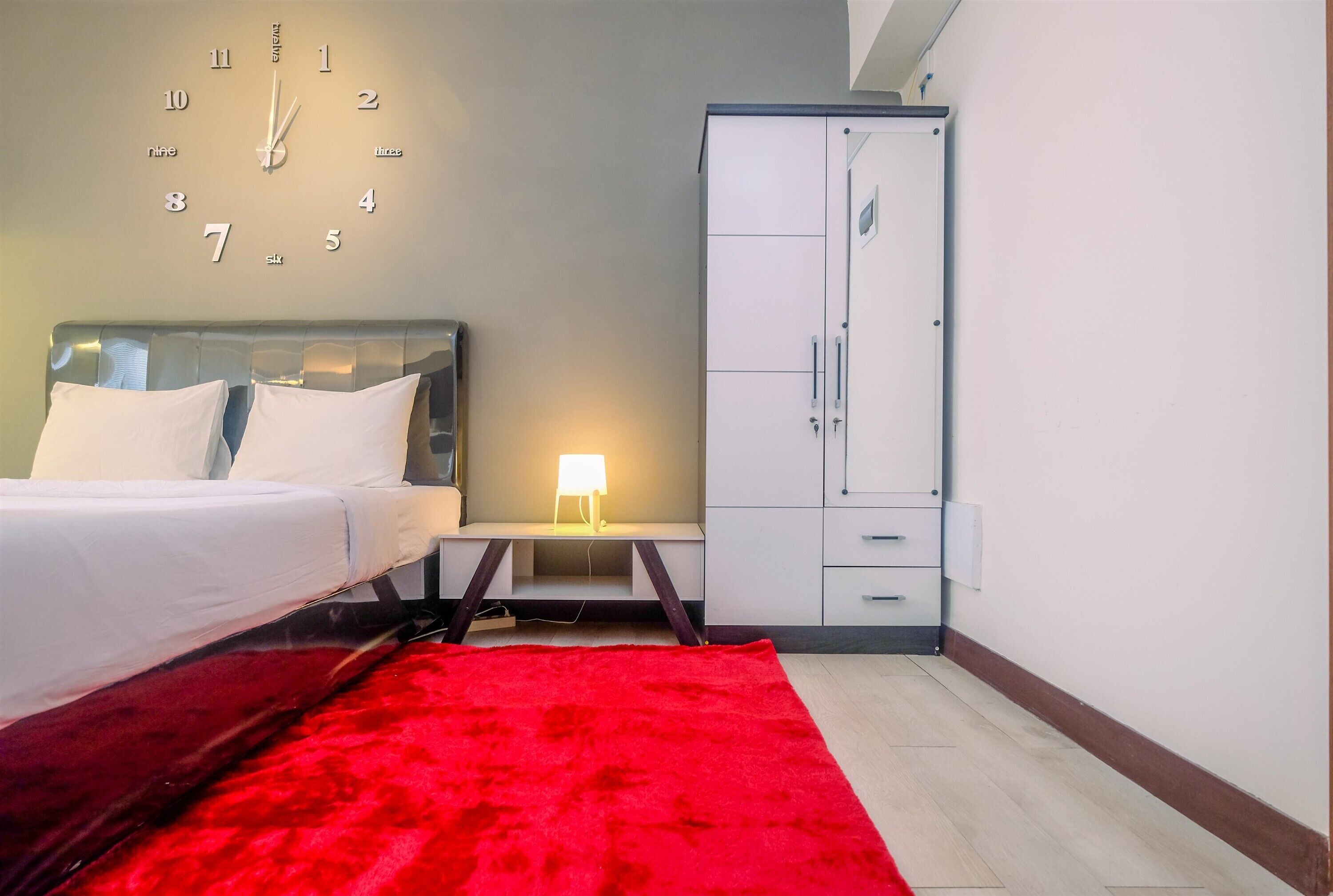 A hotel room with a bed, a nightstand with a lamp, a wardrobe, and a wall clock.
