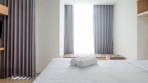 A hotel room with a large bed, a window with curtains, a wooden bench, and a TV on a stand. - Spacious Studio Room Apartment at Grand Sungkono Lagoon (Surabaya)