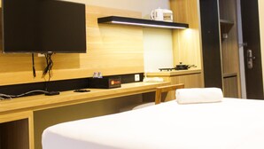 A hotel room with a bed, a flat-screen TV mounted on the wall, a wooden desk with a kettle and a tray, and a door. - Cozy and Comfortable Studio (No Kitchen) Apartment at Mustika Golf Residence (Cikarang)