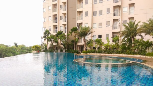 A swimming pool with a tiled edge, surrounded by palm trees and a multi-story building in the background. - Cozy and Comfortable Studio (No Kitchen) Apartment at Mustika Golf Residence (Cikarang)