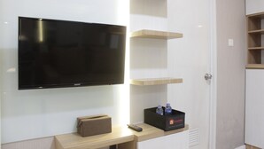 A modern hotel room with a flat-screen TV mounted on the wall, a wooden console table with a remote control, a minibar, and built-in shelving. - Favorable 1BR Apartment at Parahyangan Residence near UNPAR (Bandung)