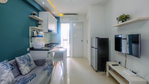 A modern kitchen with white cabinets, a built-in stove, and a refrigerator. There is a dining area with a white table and chairs, and a sofa with a leaf-patterned cover. A flat-screen TV is mounted on the wall, and a small plant is on a shelf above it. - Clean and Simply Cozy 2BR Bassura City Apartment (Jakarta)