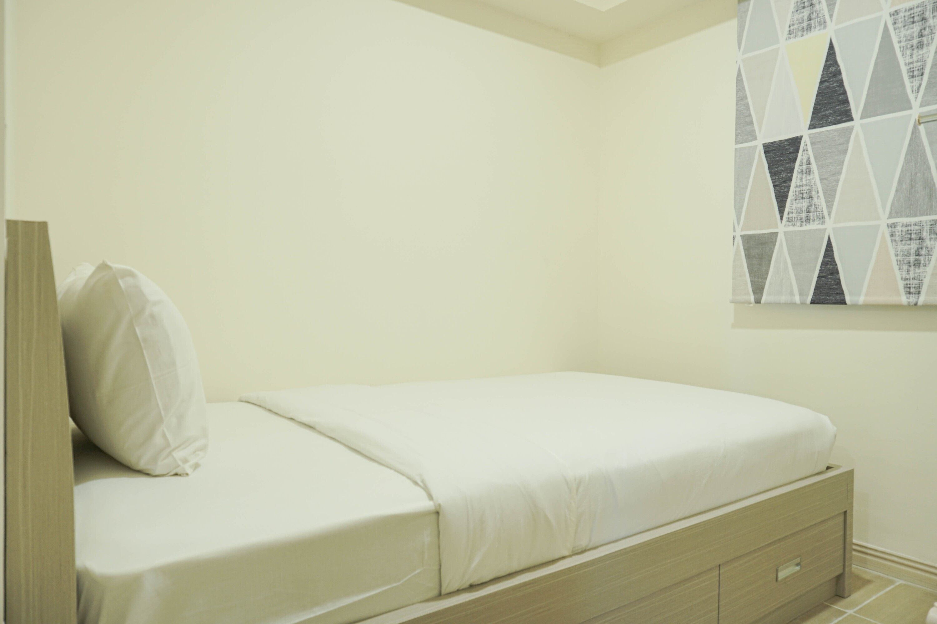 Una cama individual con estructura y cabecera de madera, un cajón de noche y una ventana con diseño geométrico instalada en la pared.
