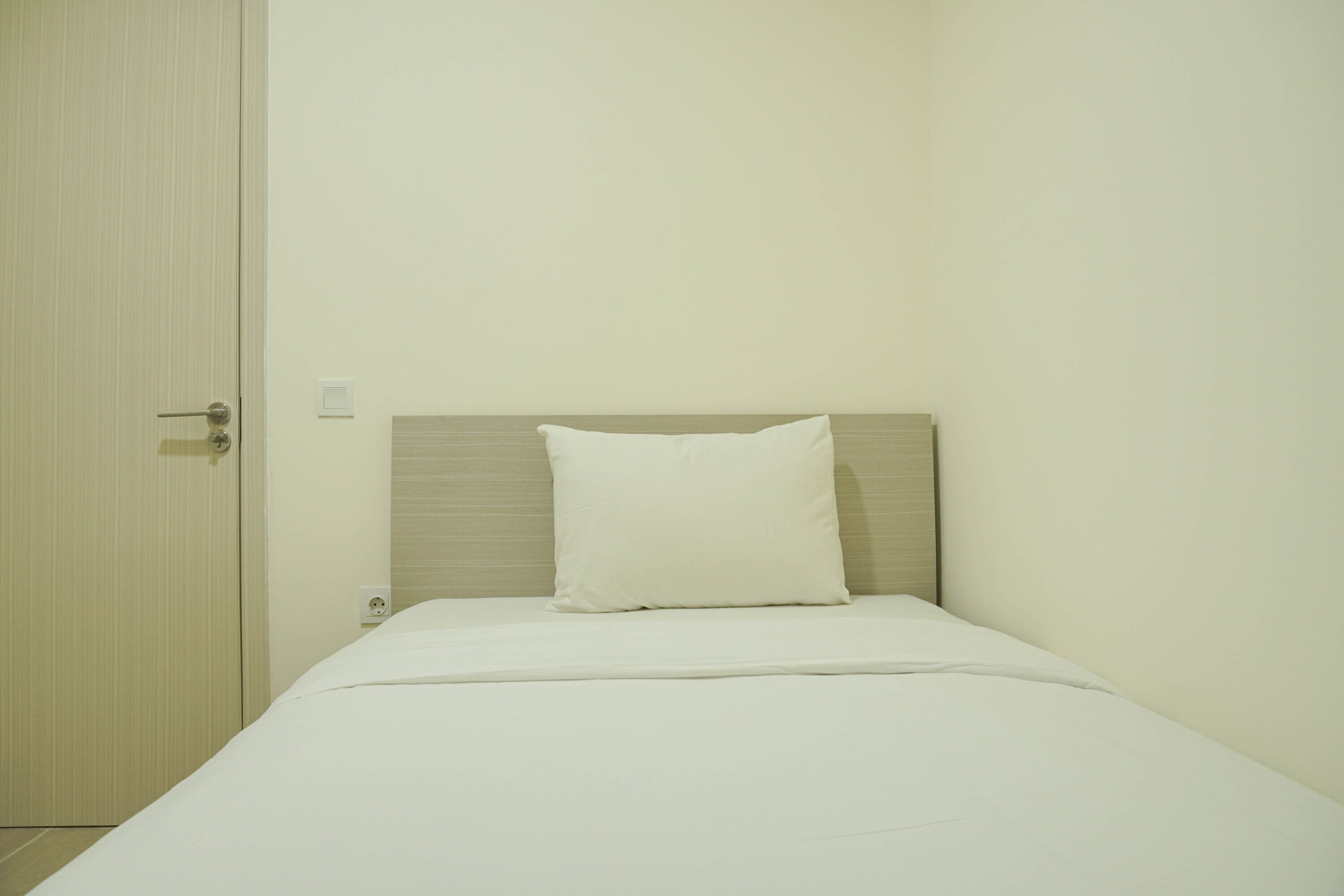 A single bed with a wooden headboard and a white pillow in a room with plain walls and a closed door.
