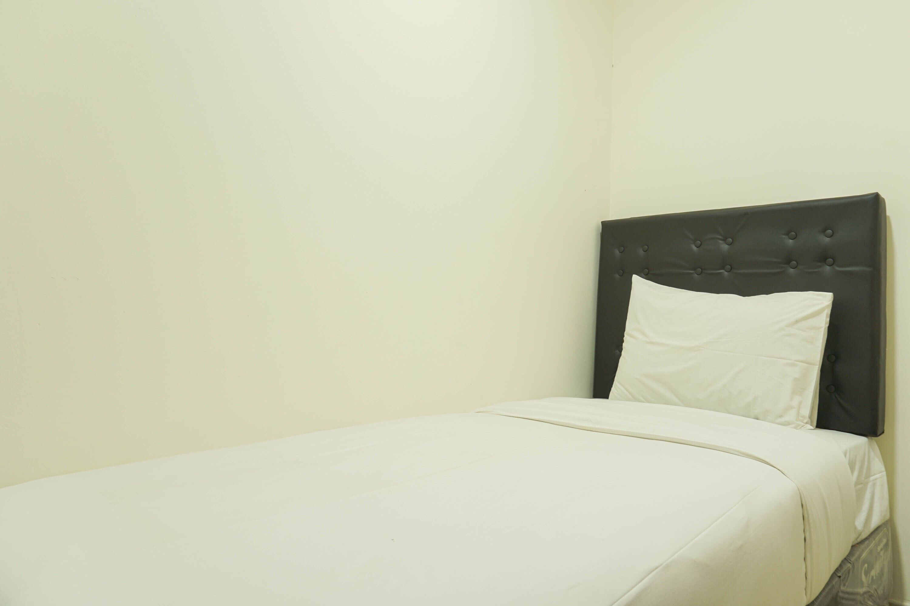 Un letto singolo con una testiera scura e un cuscino bianco in una stanza dalle pareti spoglie.