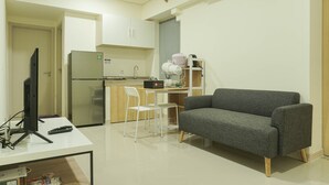 Un espace de vie compact comprenant un coin cuisine, un canapé gris, une chaise blanche et un téléviseur posé sur un bureau.