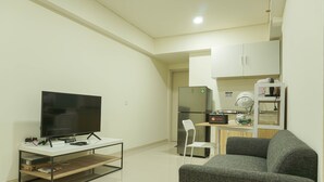 Un espace de vie compact comprenant un téléviseur à écran plat posé sur un meuble TV blanc, un canapé gris, une petite table à manger avec une chaise, un réfrigérateur et un four à micro-ondes.