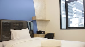 A hotel room with a bed, a nightstand, a chair, a small table with a radio, and a window with a view of a building.