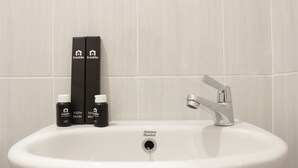 A bathroom sink with a white basin, a chrome faucet, and two black toothbrushes and toothpaste boxes labeled 'travelio' against a tiled wall. - Zest Studio Semi Apartment at The Lodge Paskal near BINUS University (Bandung)