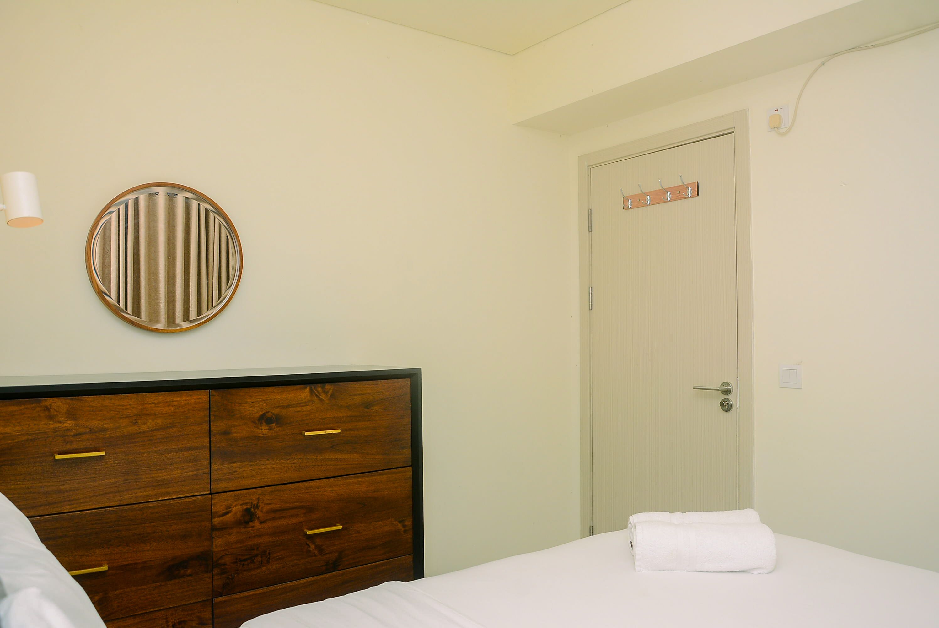 A bedroom with a wooden dresser, a round mirror, a white bed with a white towel, and a door with a coat rack.