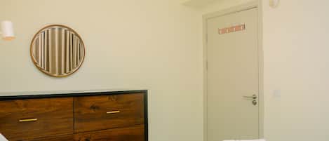 A bedroom with a wooden dresser, a round mirror, a white bed with a white towel, and a door with a coat rack.