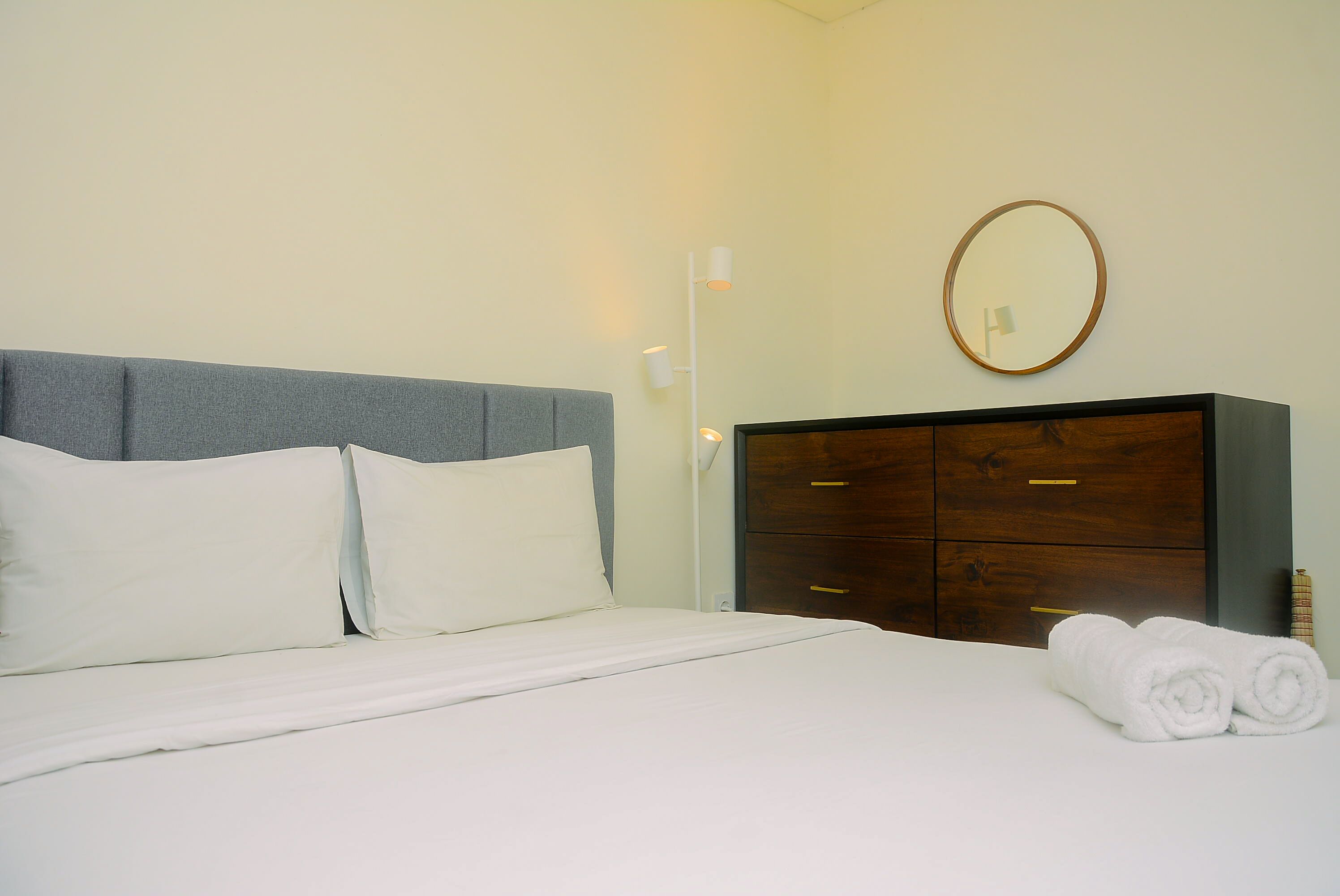 Een keurig opgemaakt bed met witte lakens, een grijze hoofdbord en een houten dressoir met lades.