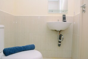 Une salle de bain avec des toilettes blanches, un mur recouvert de carreaux beiges, un lavabo blanc avec une étagère en verre au-dessus, et une serviette bleue posée sur les toilettes.