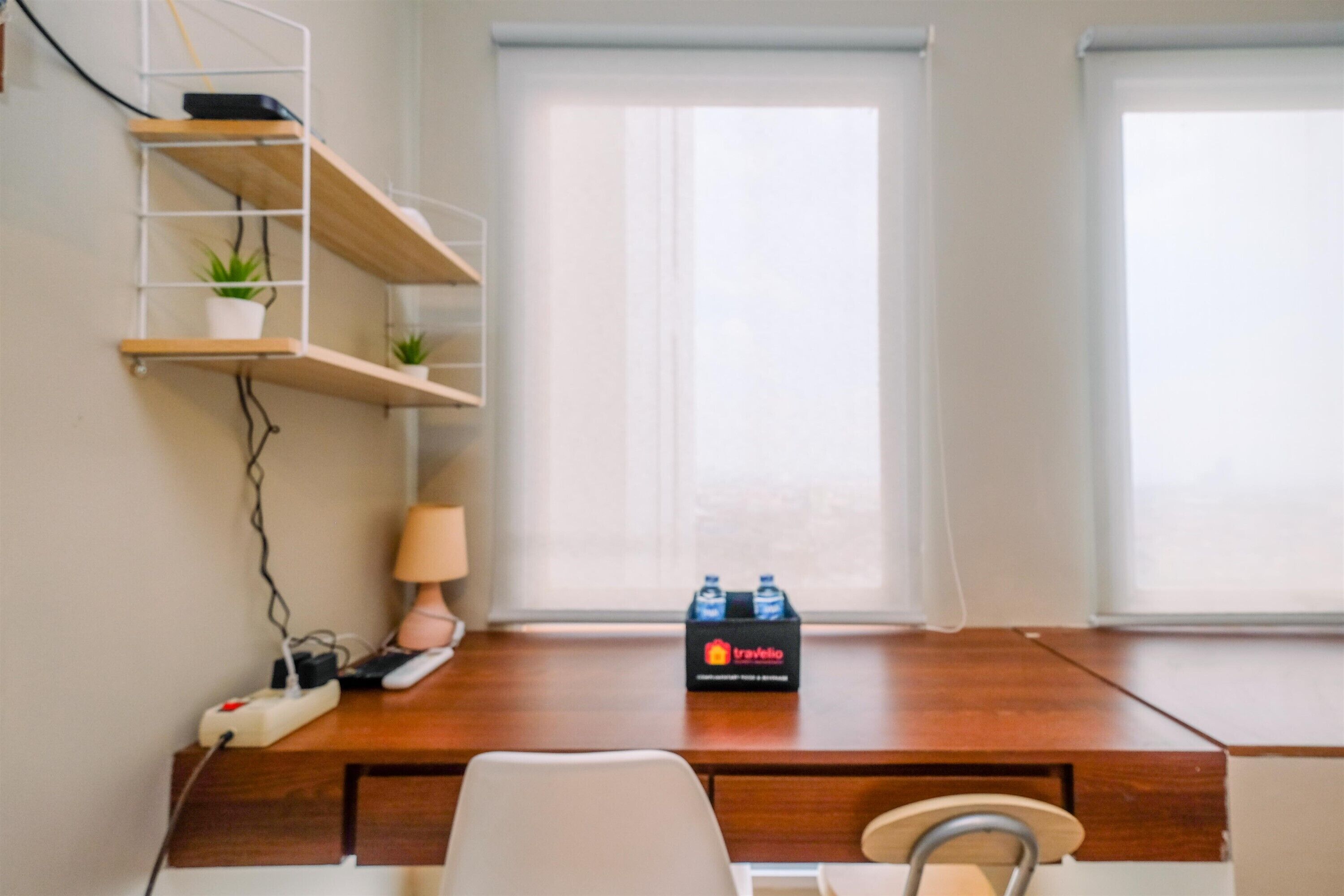 Une pièce avec un bureau en bois, une chaise, une lampe, une étagère avec des plantes et une fenêtre avec des stores.
