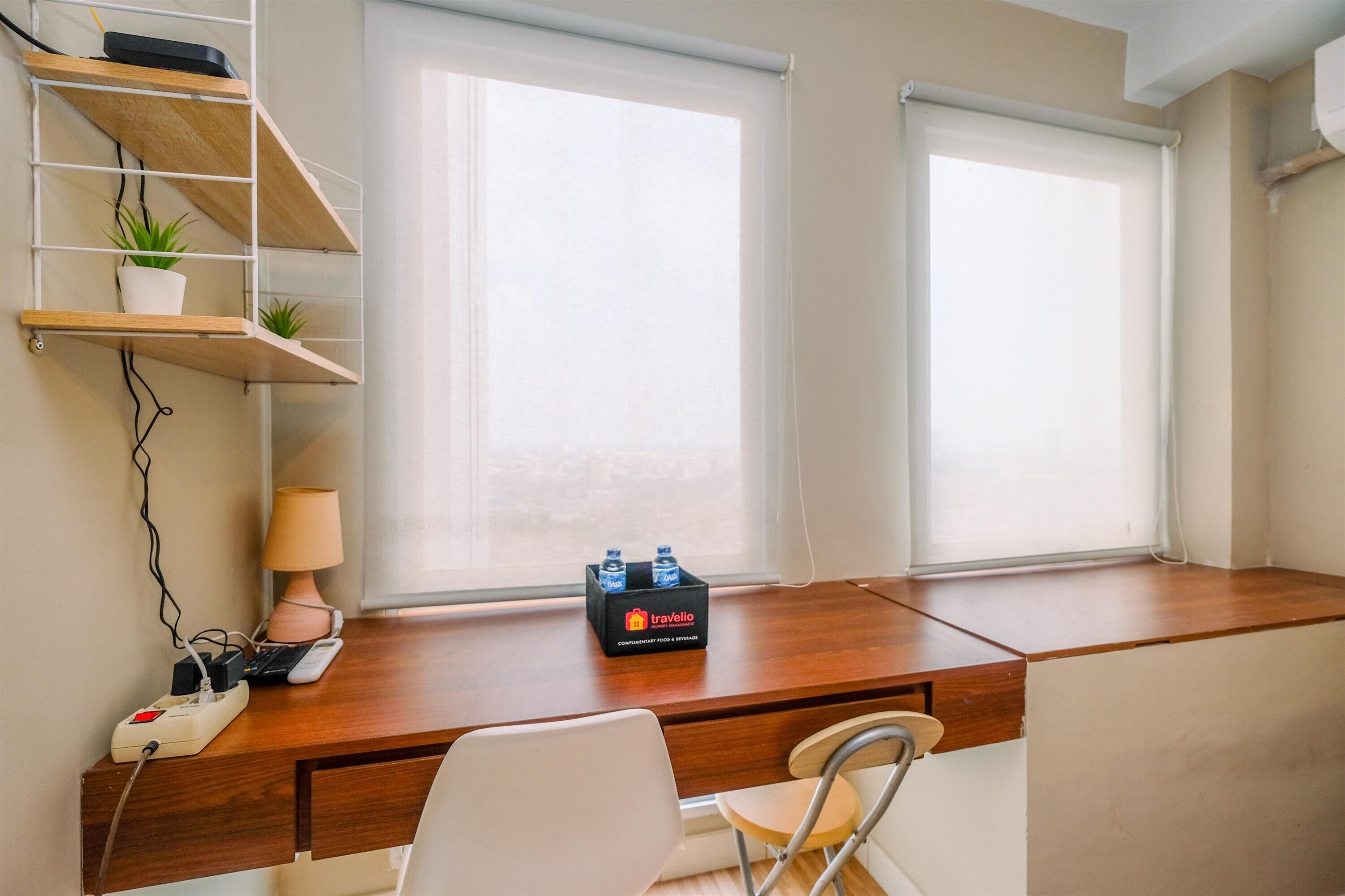 A hotel room with a wooden desk, a chair, a lamp, a power strip, a plant, and a minibar.
