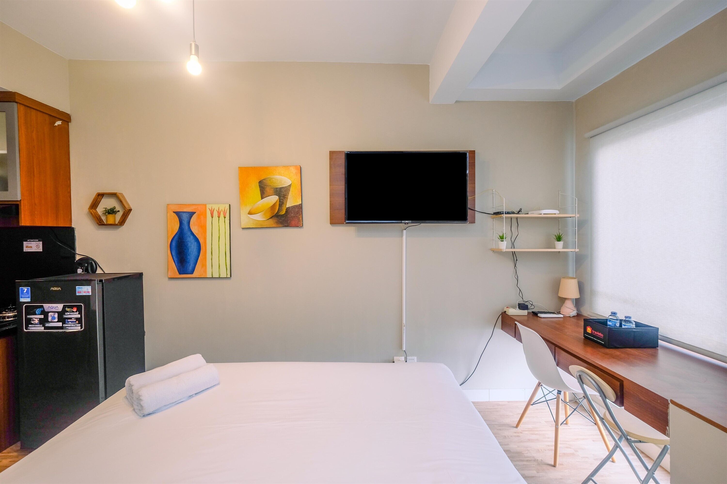A hotel room with a bed, a desk with a chair, a television, a refrigerator, and artwork on the walls.