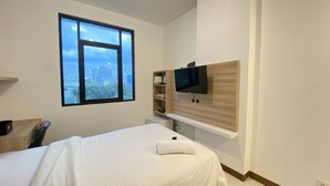 Habitación de hotel moderna con una cama grande, un televisor montado en un panel de madera, un ventanal con vistas a la ciudad y un estante pequeño con artículos de aseo.