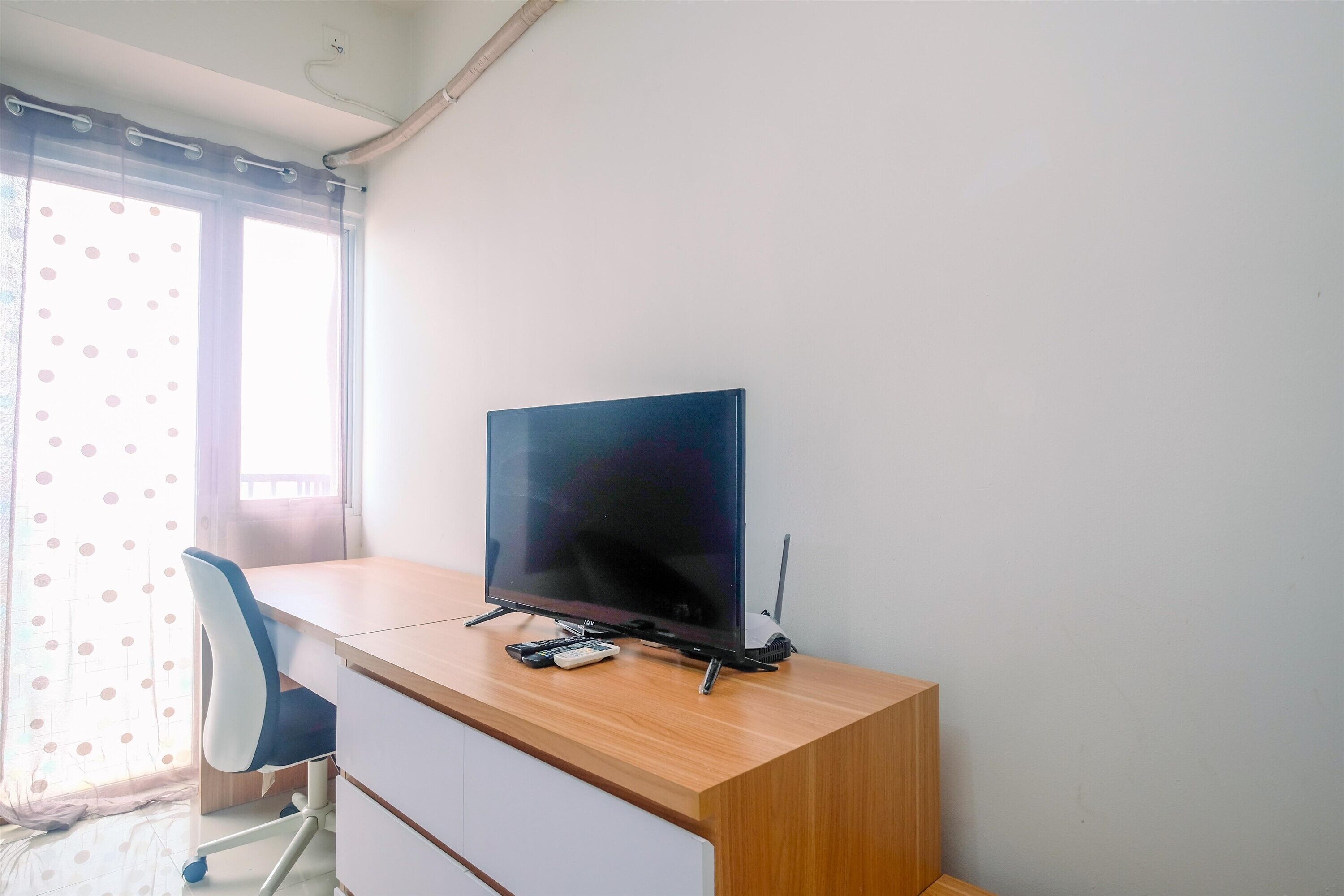 A room with a wooden desk, a chair, a television, and a window with curtains.