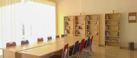 A library with a long table and chairs, bookshelves, and a trash can.