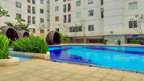 A swimming pool with a tiled deck, surrounded by greenery and a multi-story building with balconies. - Spacious and Comfort 2BR Bassura City Apartment near Mall (Jakarta)