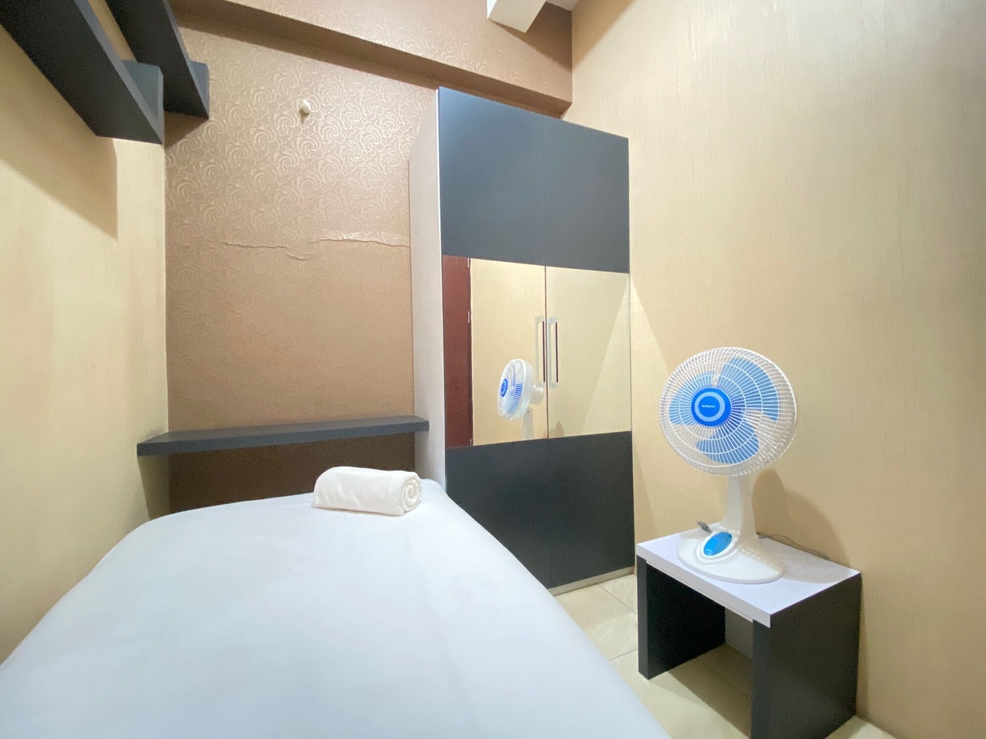 Une chambre d’hôtel avec un lit, une table de chevet équipée d’un ventilateur, une armoire et une petite étagère.