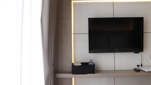 A modern hotel room with a flat-screen TV mounted on a wall, a small black shelf with a remote control, and a white laptop on a wooden shelf. - Luxurious Studio Room @ Grand Asia Afrika Apartment (Bandung)