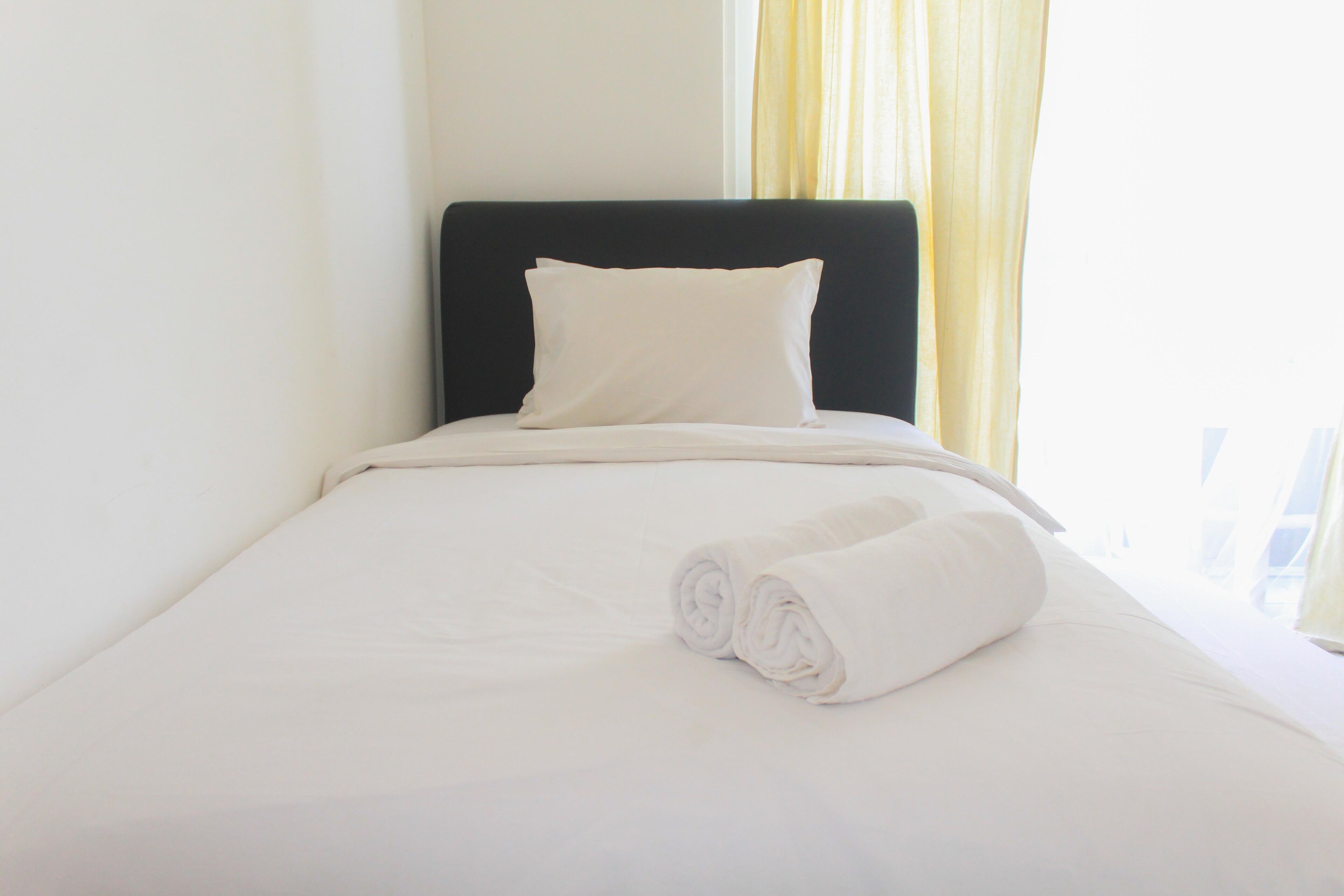 A neatly made bed with white linens and a rolled towel, a black headboard, and a window with yellow curtains.