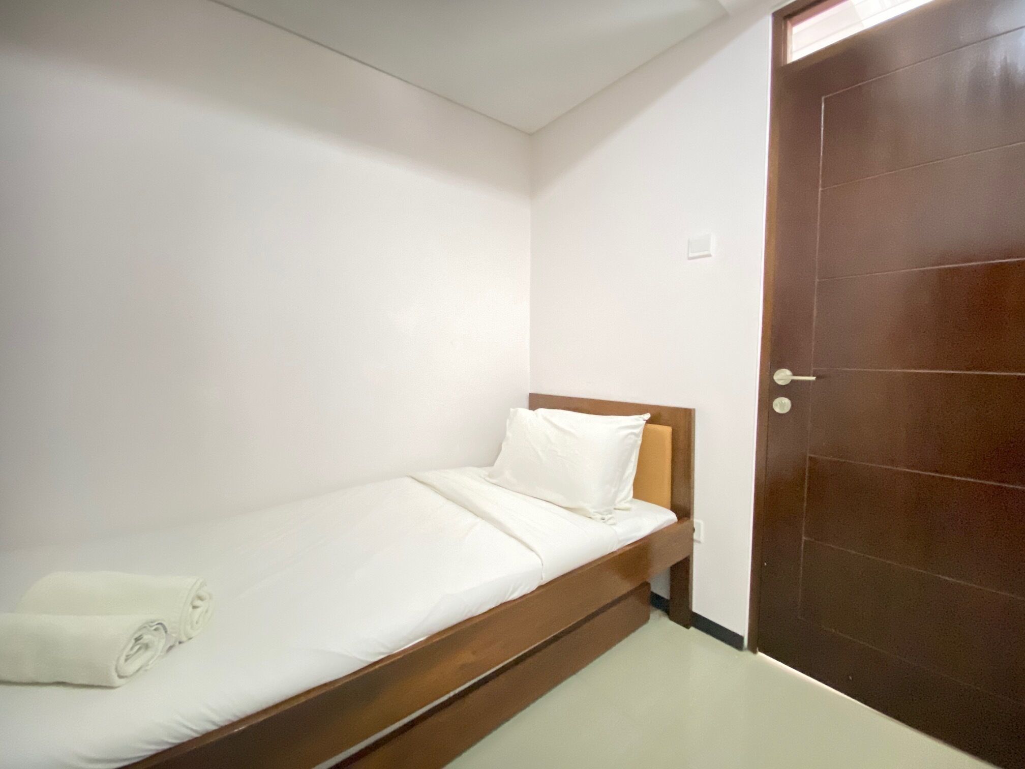 Una habitación pequeña con una cama individual, cabecera de madera, ropa de cama blanca y puerta cerrada.