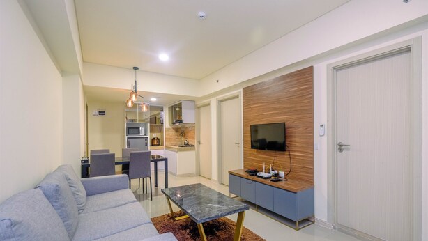 Un moderno salón con un sofá gris, una mesa de centro de vidrio y un televisor montado en una pared de madera. En el fondo, se observa un comedor con una mesa y sillas.