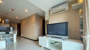 A modern living room with a flat-screen TV on a white entertainment unit, a kitchen area in the background, and a large window with curtains.
