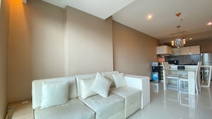 A modern living room with a white sofa, a dining area with a glass table and chairs, and a kitchen in the background.