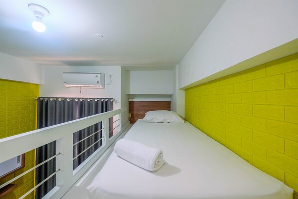 A single-bed room with a white bed, a yellow brick wall, a white ceiling light, and a window with curtains.