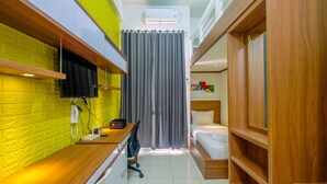 A compact hotel room with a bed, a desk with a chair, a small table, a telephone, and a wall-mounted TV. - Comfort Studio with Bunk Bed at Dave Apartment (Depok)