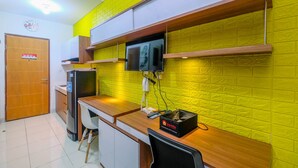 A compact hotel room with a wooden desk, a television mounted on a brick-patterned wall, a refrigerator, and a small cabinet. - Comfort Studio with Bunk Bed at Dave Apartment (Depok)