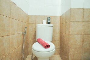A small, tiled bathroom with a toilet, a shower head, and a roll of toilet paper on the tank.
