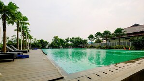 A swimming pool with a wooden deck and lounge chairs, surrounded by palm trees and a building with a tiled roof. - Gorgeous and Strategic Studio Apartment at Capitol Park Residence (Jakarta)