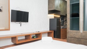 Una habitación de hotel con una cama, un televisor montado en la pared, un banco de madera, un estante pequeño y una zona de cocina con un refrigerador y armarios.
