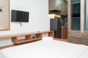 Una habitación de hotel con una cama, un televisor montado en la pared, un banco de madera, un estante pequeño y una zona de cocina con un refrigerador y armarios.