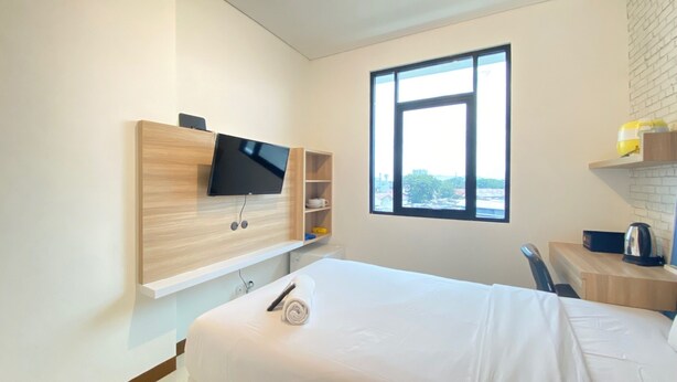 A modern hotel room with a bed, a TV mounted on a wooden shelf, a window with a city view, and a small desk with a kettle.
