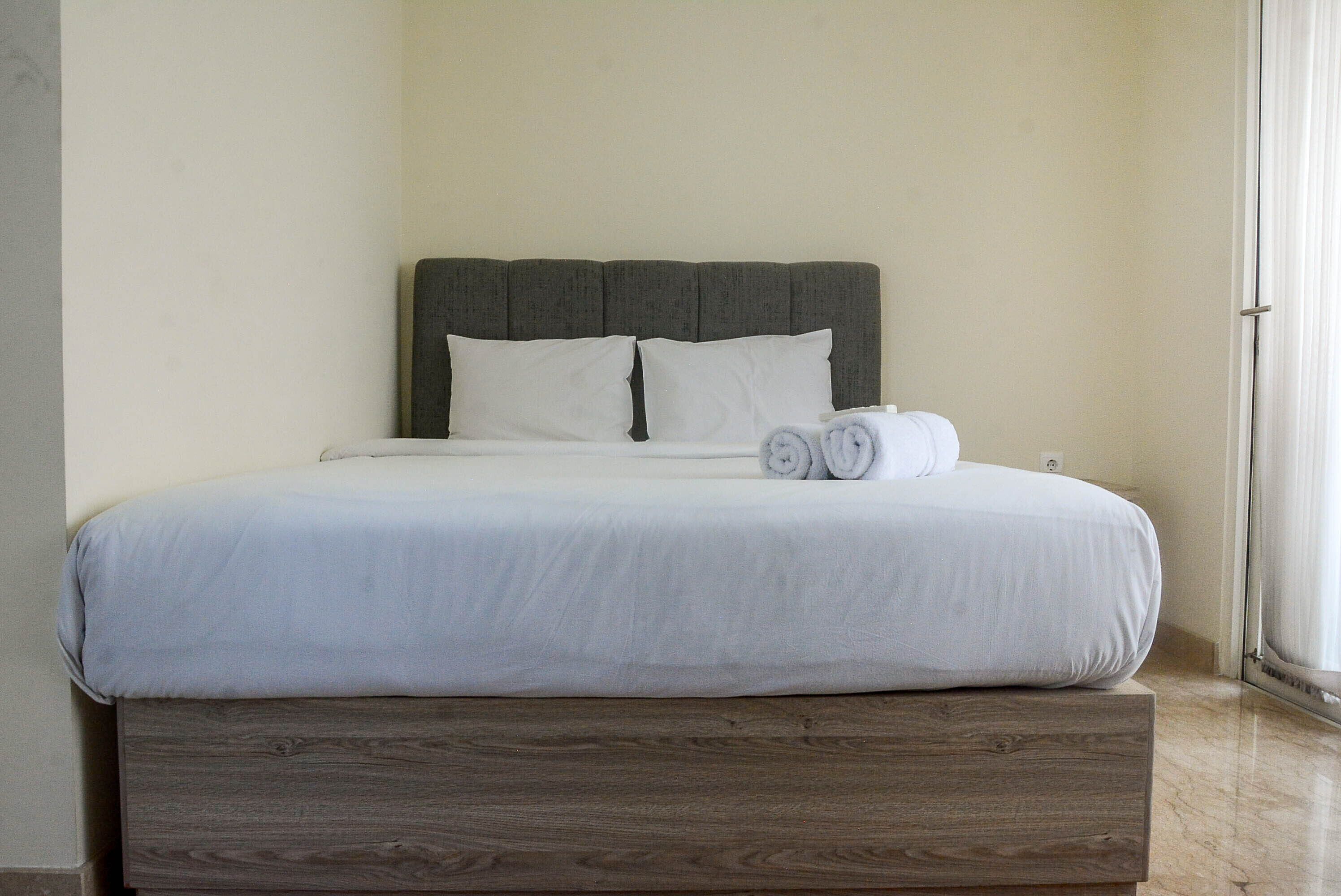 Una cama bien tendida con sábanas blancas y dos toallas enrolladas, un cabecero tapizado gris y una base de madera.