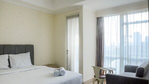 A hotel room with a bed, a chair, a small table, and a view of the city through large windows. - Great Location Studio at Menteng Park Apartment (Jakarta)