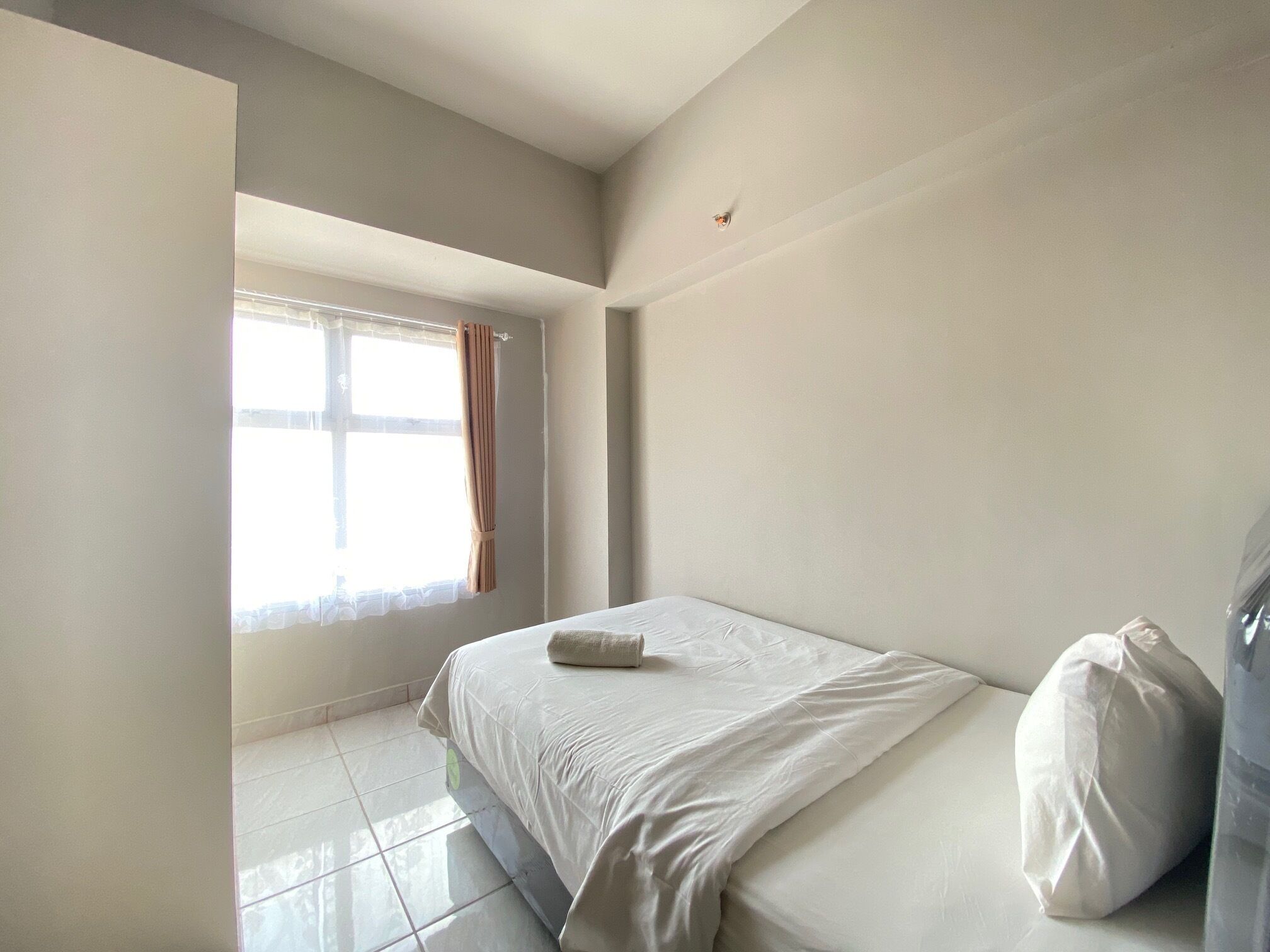 A bedroom with a bed, a window with curtains, a tiled floor, and a small wall-mounted light fixture.