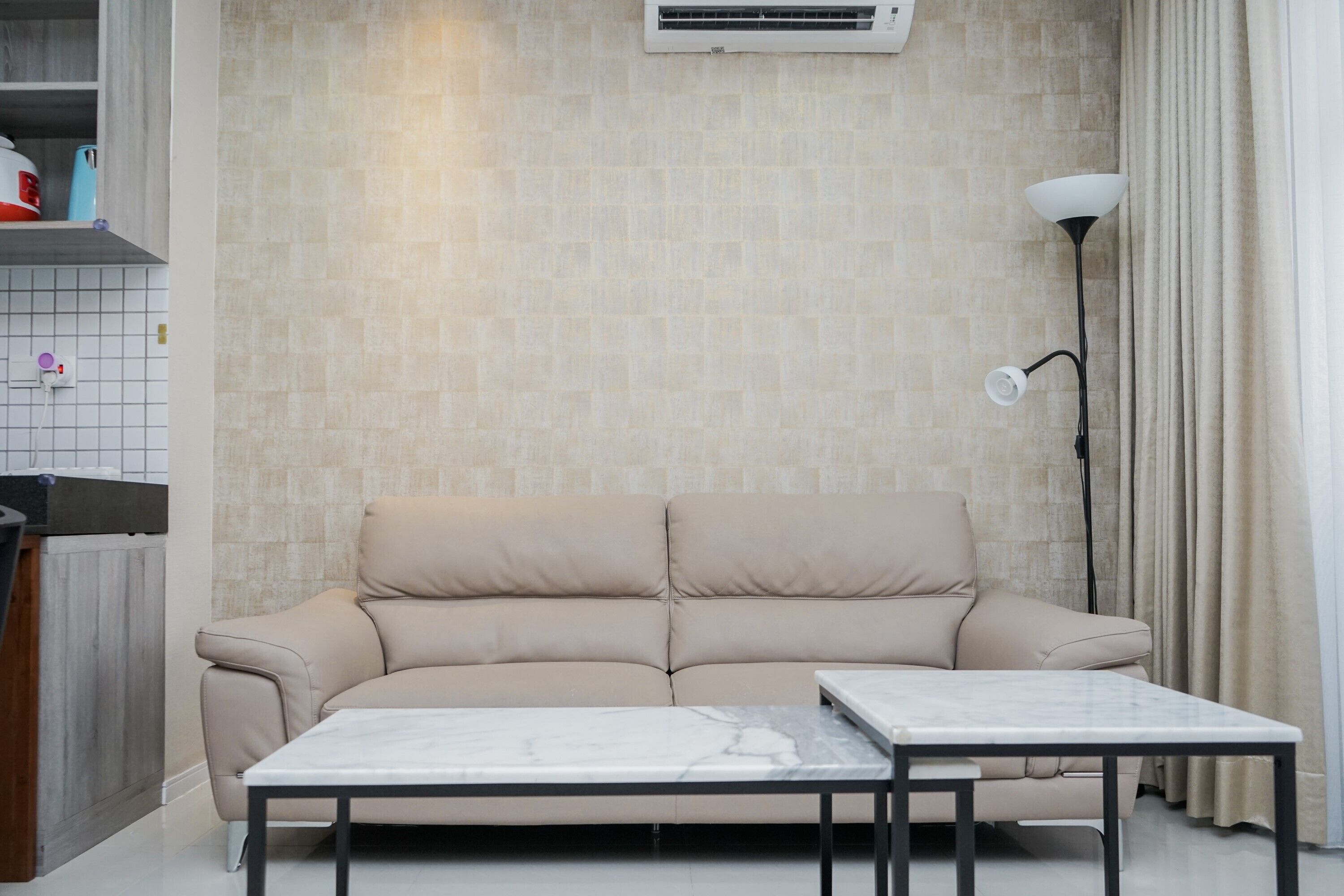 En stue med en beige sofa, et marmorbord, en gulvlampe og en flisevæg med et klimaanlæg.