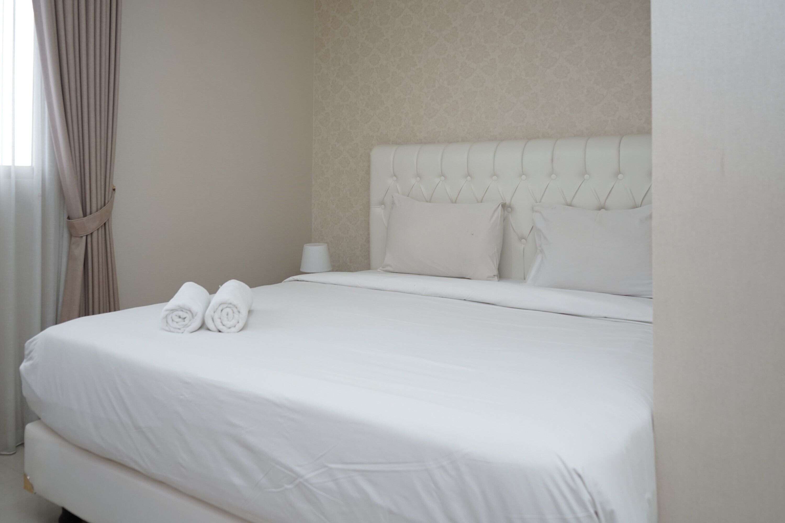 A neatly made bed with white linens and two rolled towels, a bedside table with a lamp, and a headboard with tufting.