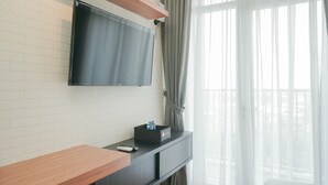Habitación de hotel moderna con una televisión de pantalla plana montada en una pared de ladrillo, una mesita consola de madera, una pequeña caja negra y vistas a un balcón a través de cortinas de tela fina.